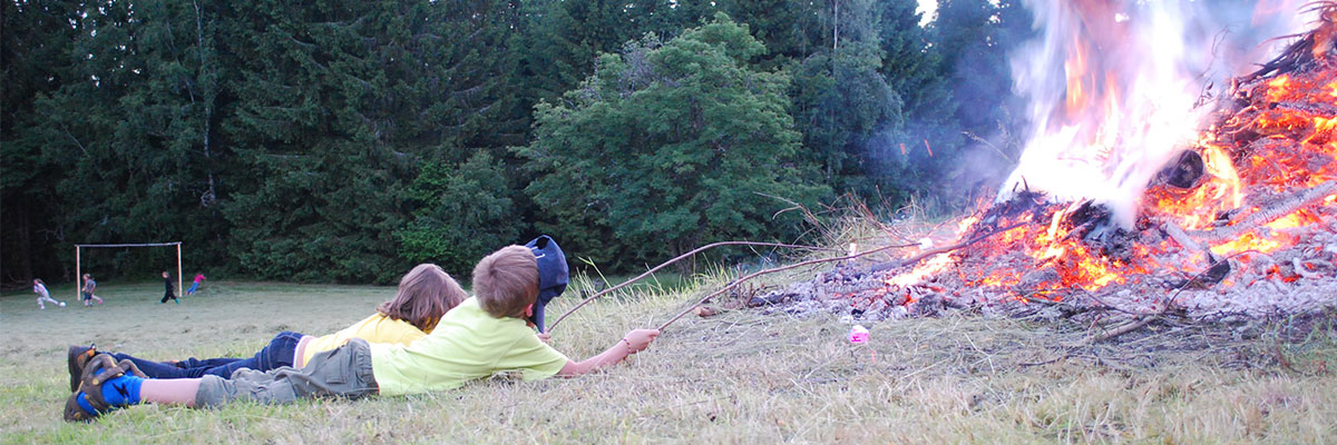 Zwei Kinder liegen in der Wiese und halten lange Stöcke in ein Feuer. Der eine Junge schützt sein Gesicht mit einer Kappe vor der Hitze der Flammen.